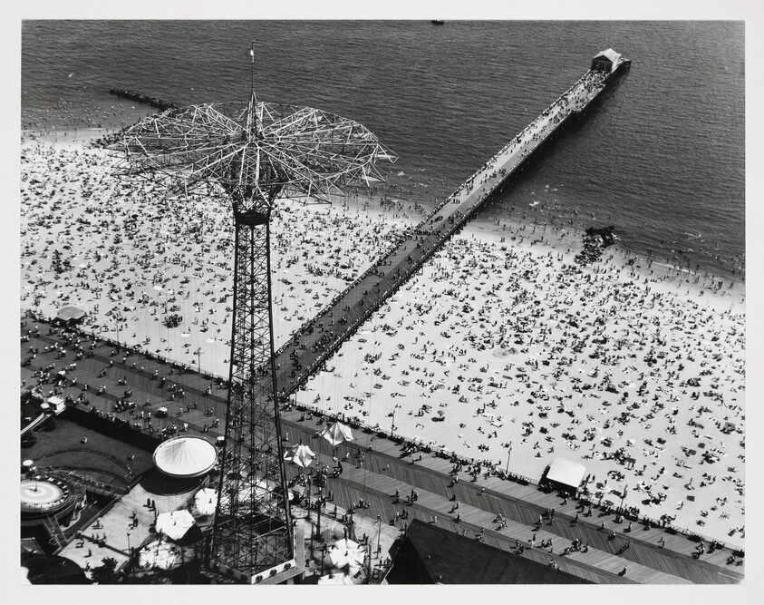 A tall parachute-jump tower rises beside a crowded beach with a long pier stretching into the ocean.