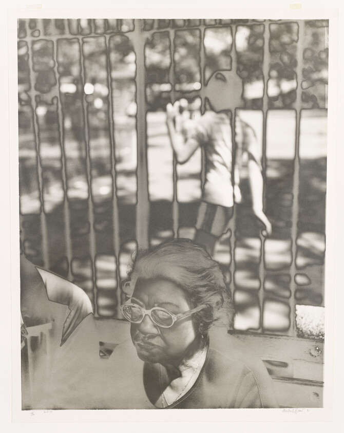 An elderly woman with glasses sits in front of a metal fence while a child peers through it.
