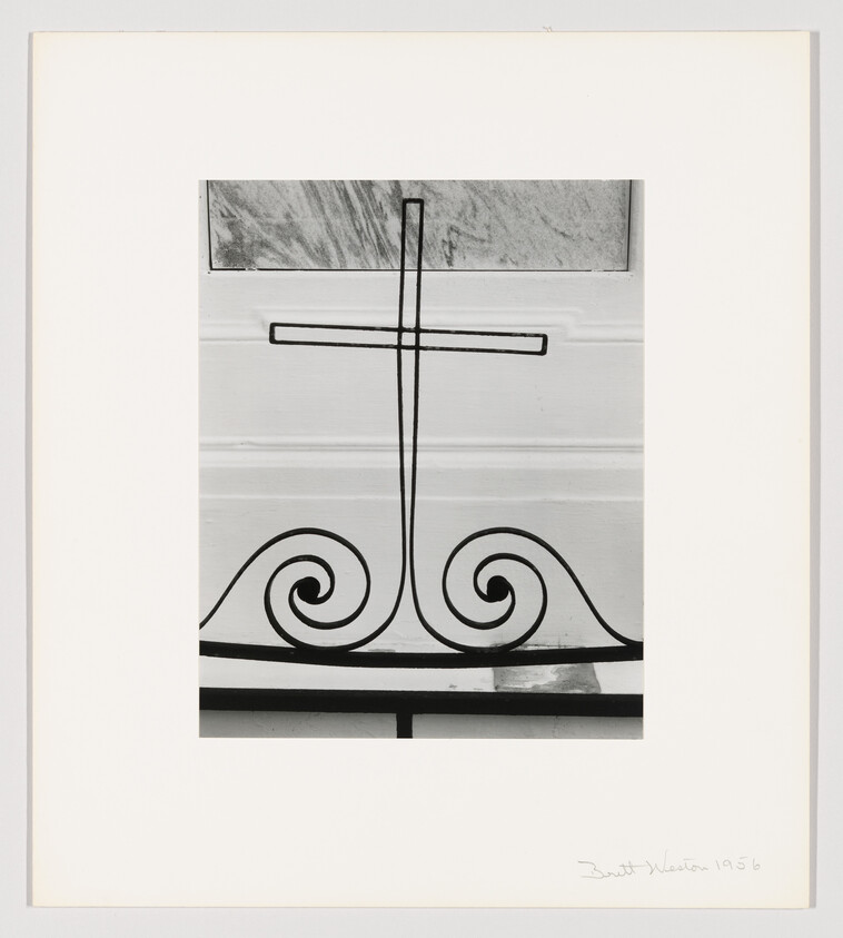 Wrought-iron cross with two symmetrical scrolls mounted on a white paneled wall.