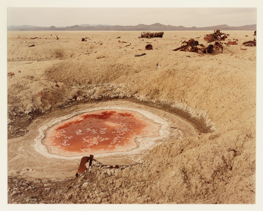 A shallow crater with a red salt pool in a dry desert surrounded by rusty vehicle wrecks.