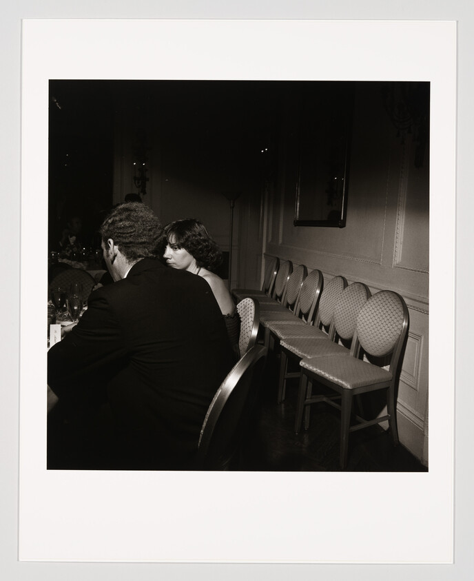 A woman sitting at a dim banquet table looks over her shoulder while empty chairs line the wall.