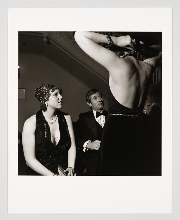 Two seated guests watch a standing woman adjust a necklace on her backless evening gown.