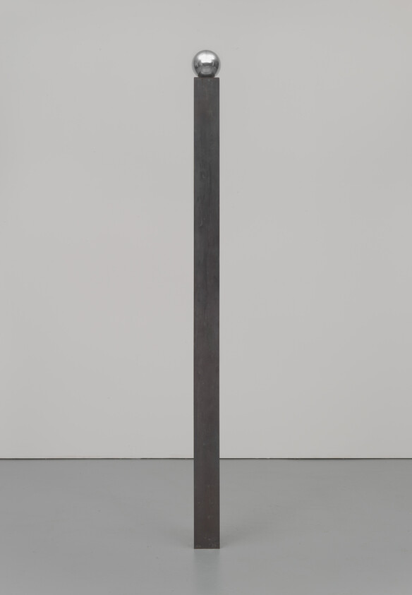 A thin, dark bronze column standing alone with a balanced steel ball resting on top of the exact diameter