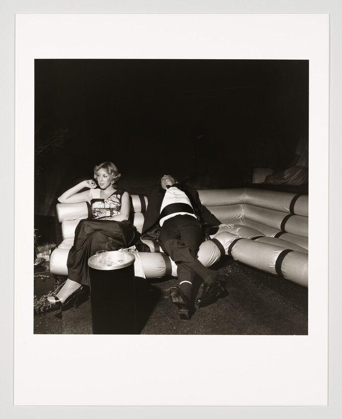 A formally dressed woman sits pensively while a man in a suit slumps asleep on an inflatable couch.