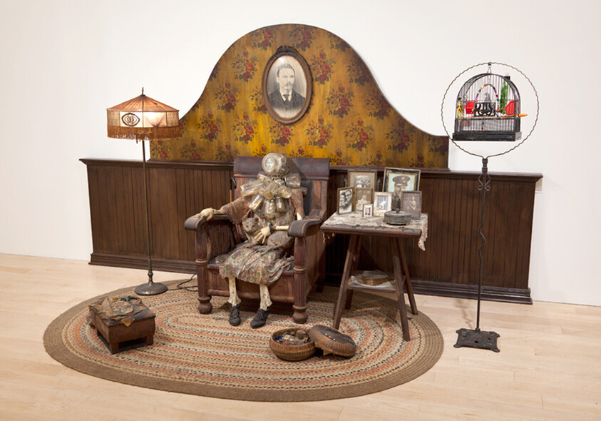 A seated cloth mannequin wearing layered clothing sits in an armchair surrounded by family photos and a birdcage.