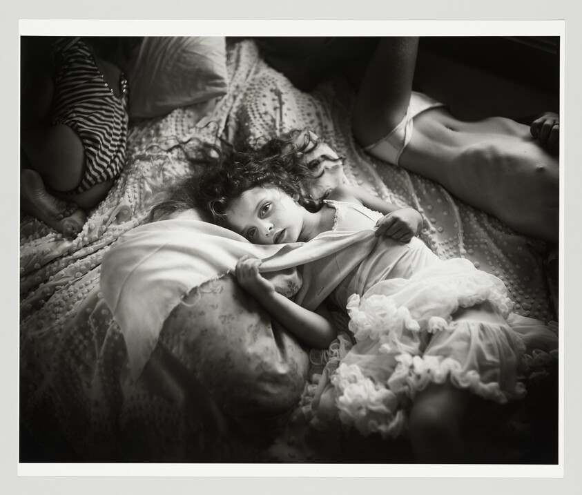 A young girl lies on a bed holding a blanket and gazing directly at the camera.