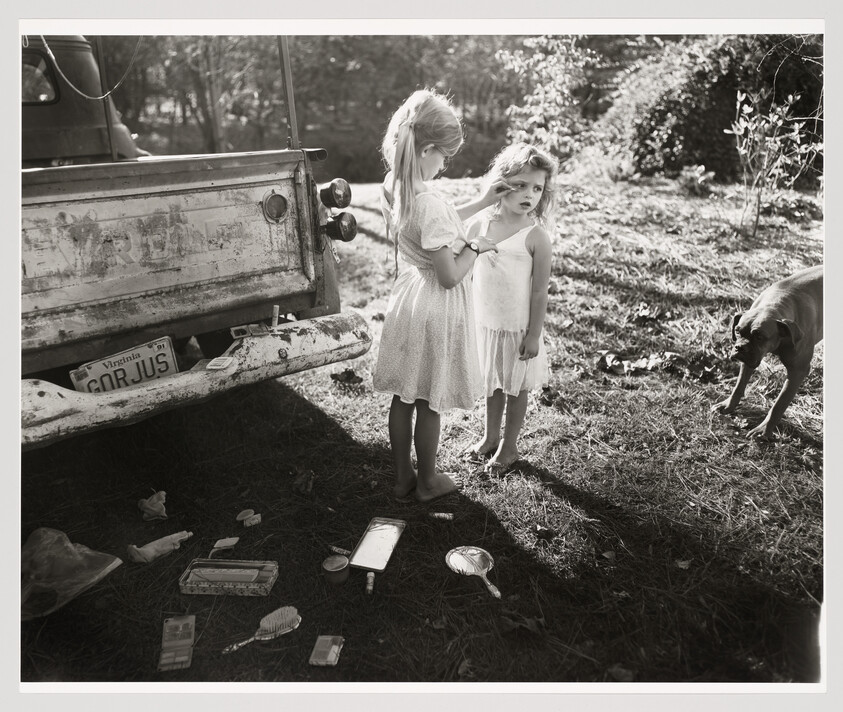 Two young girls stand by an old pickup while one brushes the other's hair amid scattered makeup.