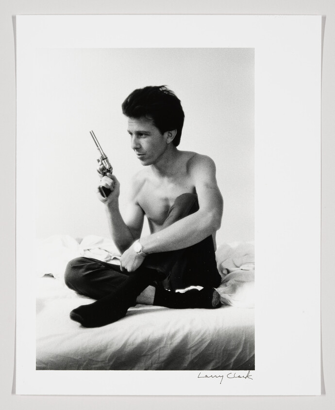 A shirtless young man sits cross-legged on a bed holding a revolver and looking to the side.