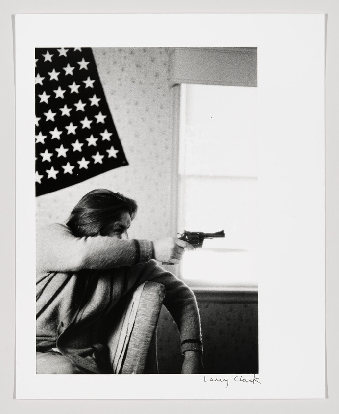 Man aiming a revolver from a chair toward a window with a US flag on the wall.