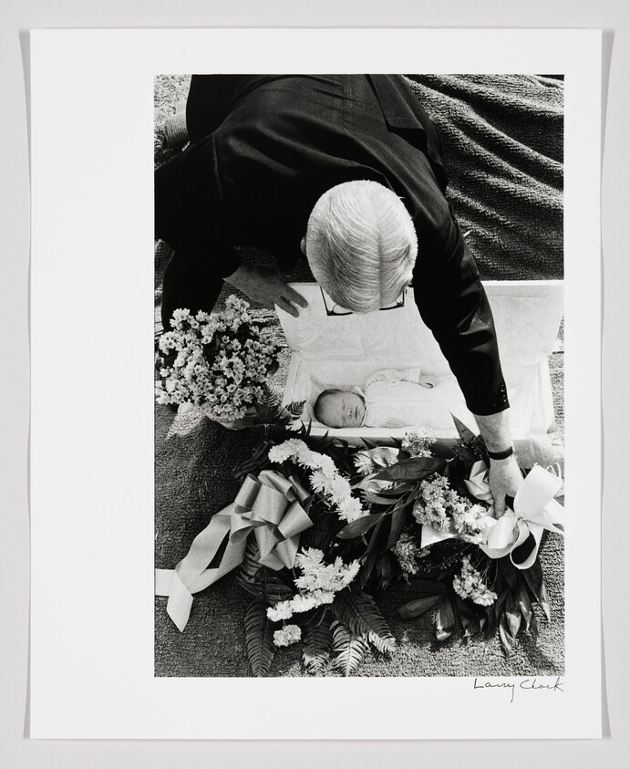An older man leans over a small open casket holding a sleeping infant, surrounded by flowers.
