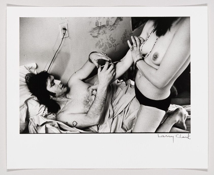 Man lying in bed holds a syringe against a woman's arm while she stands beside him topless.