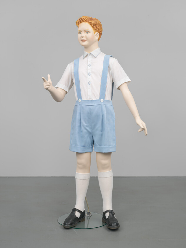 A child mannequin wearing light blue shorts with suspenders and a white shirt stands with one hand raised.