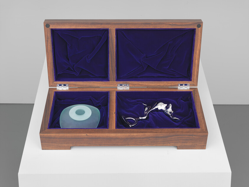 Open wooden display box with purple velvet lining holding a blue glass ring and small silver sculpture.