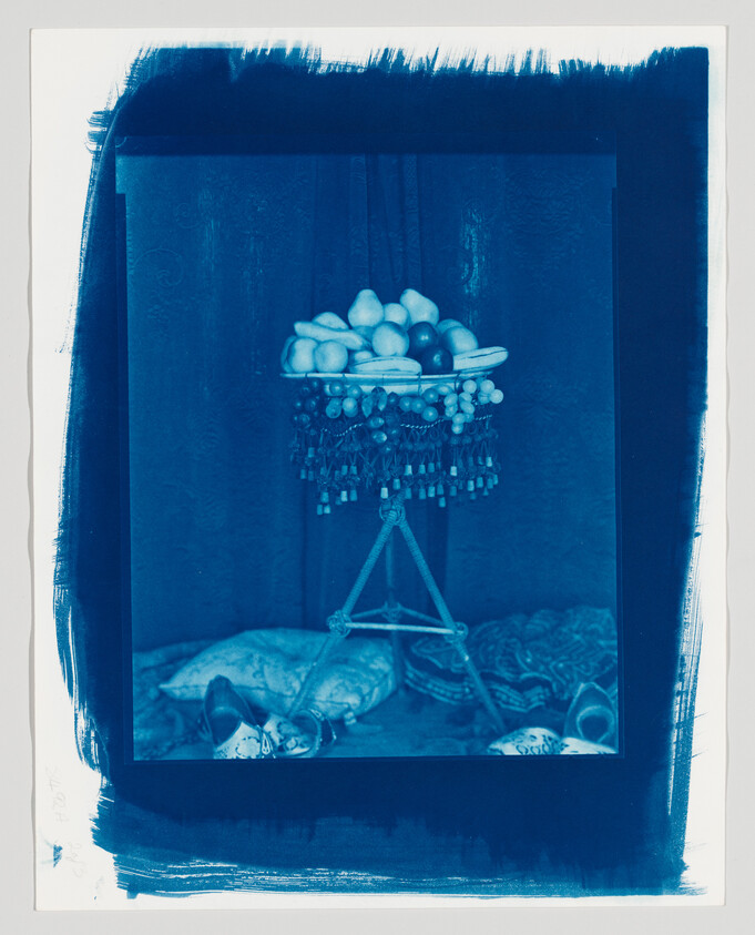 A decorative fruit-filled bowl sits on a beaded tripod stand above cushions and slippers.