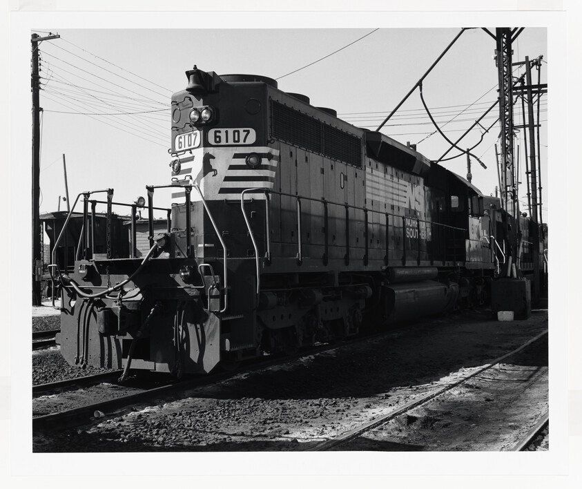 A diesel locomotive numbered 6107 idles on railroad tracks beside overhead power lines.