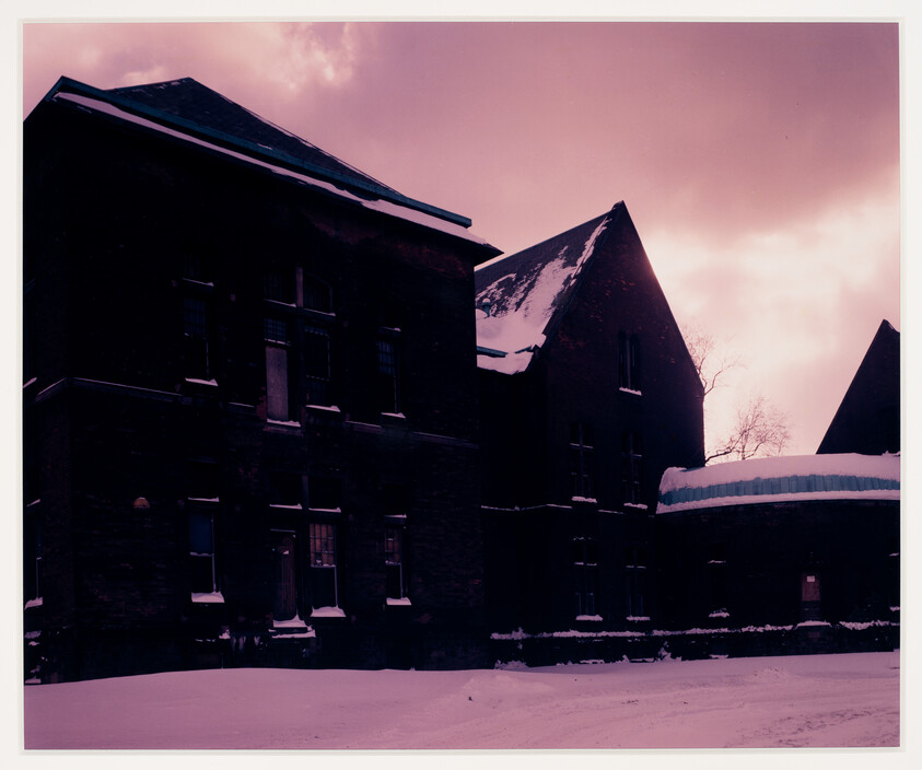 Large brick building with snow on the roof and ground under a pink sky.