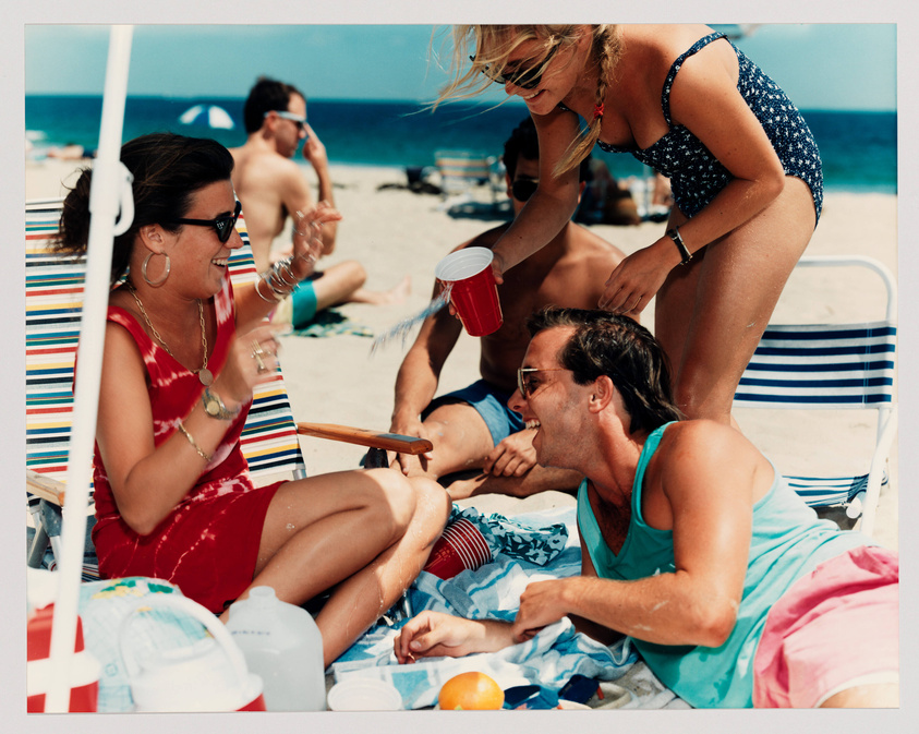 A group of people enjoying a sunny day at the beach, with one woman sitting on a beach chair laughing and holding a red cup, another woman bending over to talk to a man lying on a towel, and various beach items scattered around, including a cooler and an orange. The ocean and other beachgoers are visible in the background.