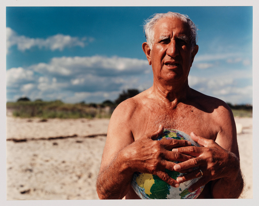 An elderly man with gray hair stands shirtless on a beach, holding a small globe in his hands close to his chest. The sky is blue with scattered clouds in the background.