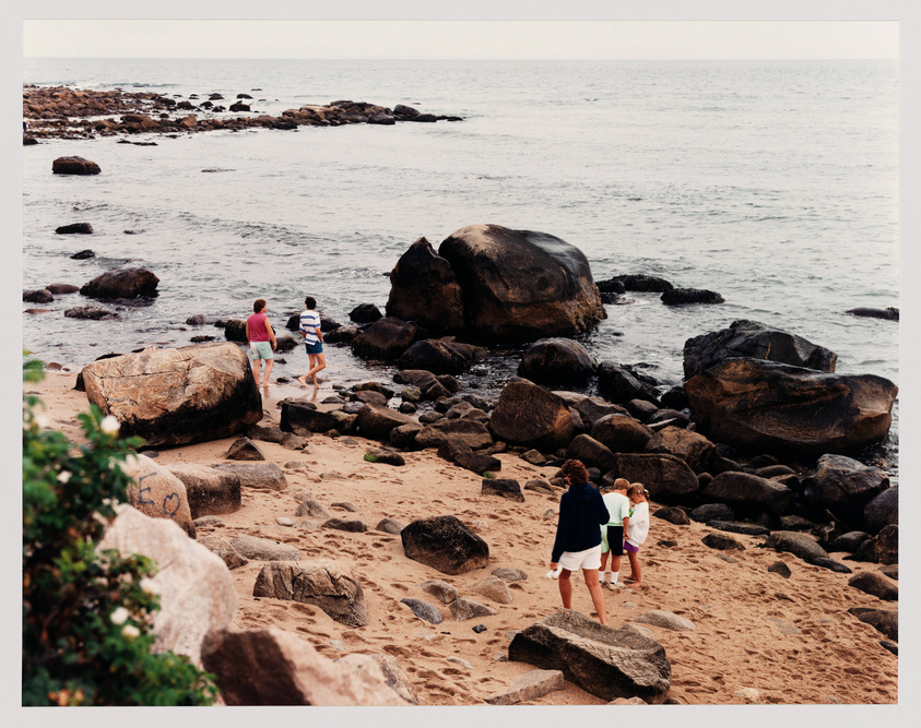 A sandy beach with scattered rocks and boulders leading to the ocean, with four people walking near the water's edge.