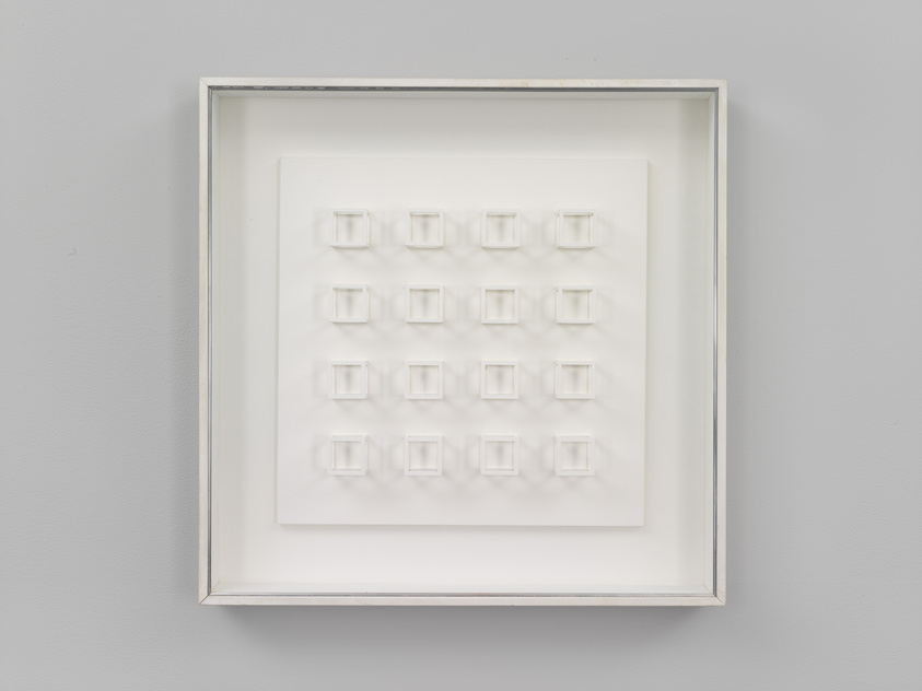 A framed three-dimensional white artwork featuring a grid of square indentations, displayed on a light gray wall.