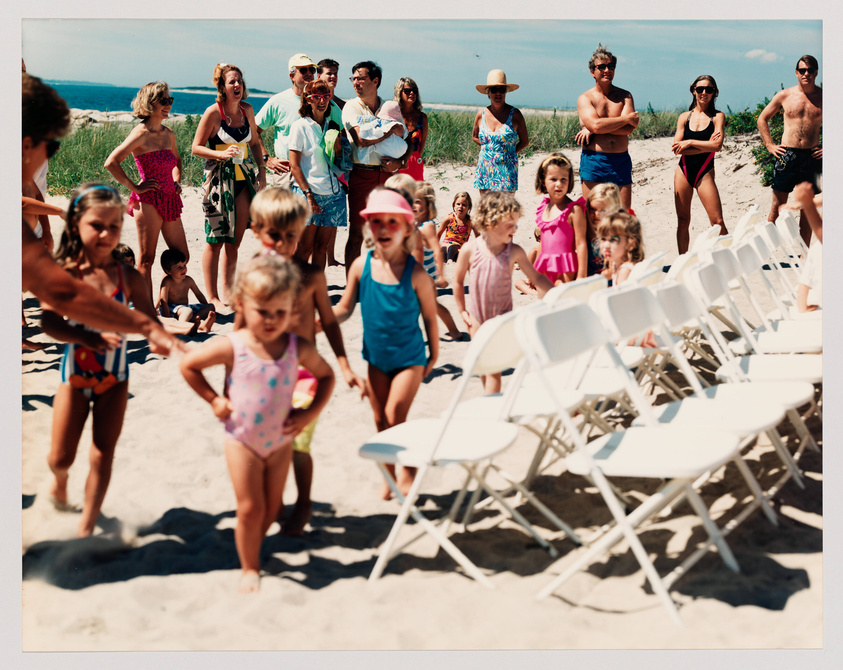 A group of people, including children and adults, are gathered on a sandy beach on a sunny day. The children, wearing colorful swimsuits, are in the foreground, some walking towards the camera, while the adults stand or sit in the background, observing. A row of white folding chairs is arranged on the right, and the blue sky and ocean are visible in the distance. Everyone appears to be enjoying a beachside event or activity.