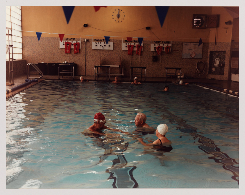 An indoor swimming pool with several swimmers, some wearing swim caps, leisurely floating or swimming. The pool area is adorned with bunting flags and signs indicating different swimming lane speeds such as "Slow," "Medium," and "Fast." A large clock on the wall shows the time, and various swimming-related notices and equipment are visible around the poolside.