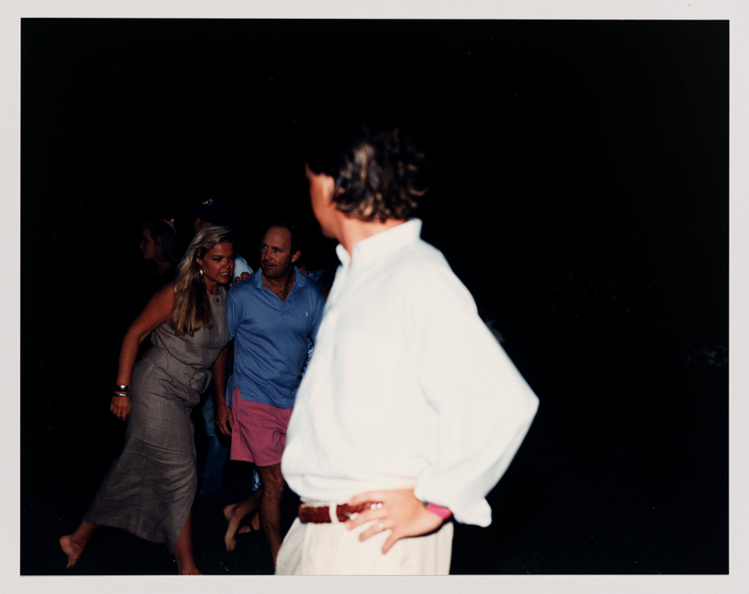 A group of people in casual evening wear gathered in a dimly lit outdoor setting, with a woman and a man in the center appearing to be in conversation, and another person's back in the foreground facing away from the camera.