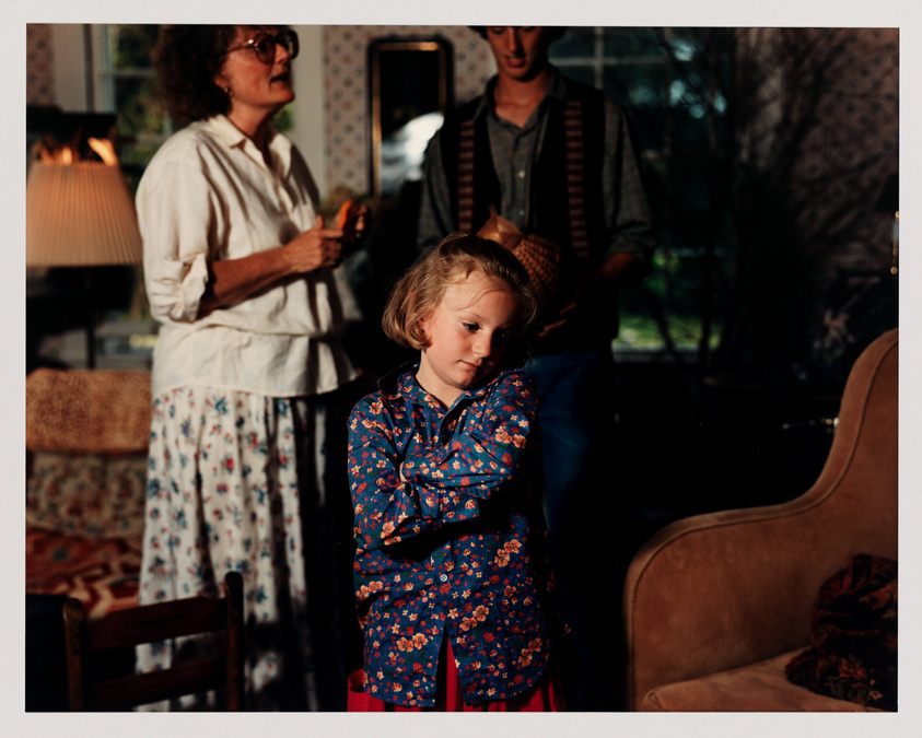 A candid photo capturing a moment in a cozy living room with vintage decor. A young child in a blue floral shirt stands in the foreground, looking to the side with a contemplative expression. Behind the child, a woman in a white blouse and floral skirt stands holding an object, looking off to the side, while a man in a striped vest and hat is partially visible in the background. The room is warmly lit, suggesting a late afternoon or evening setting.