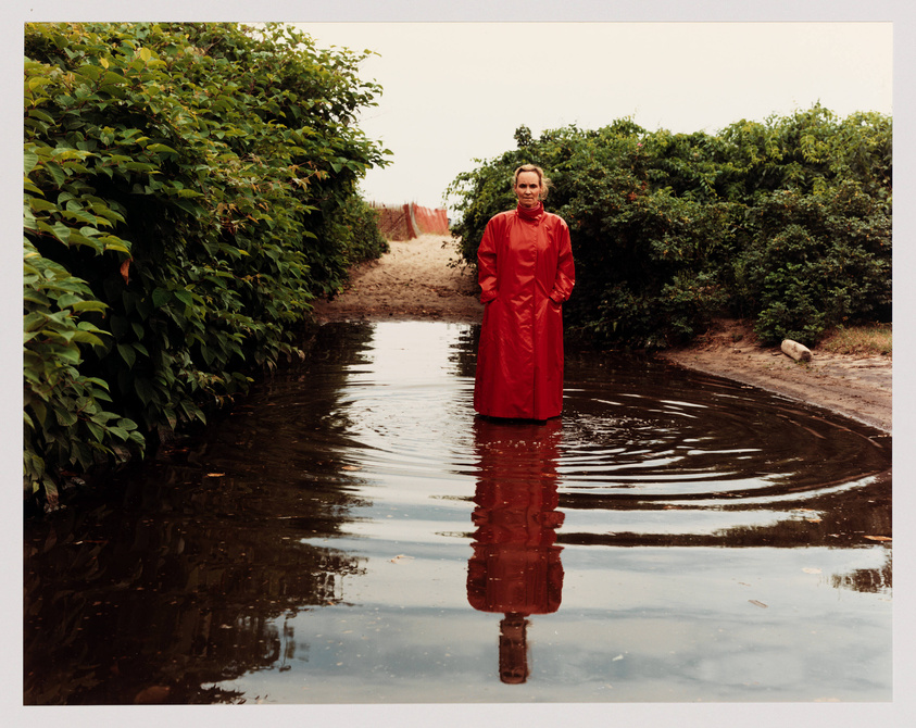 A person in a red dress stands centered in a large puddle of water on a dirt path, flanked by lush greenery, with ripples emanating from where they stand.