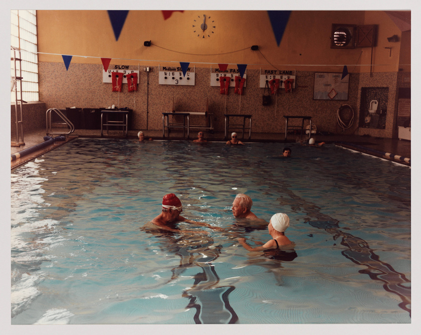 A group of elderly swimmers wearing swim caps leisurely enjoy time in an indoor swimming pool, with signs indicating different swimming speeds on the wall and various swimming-related notices and decorations.