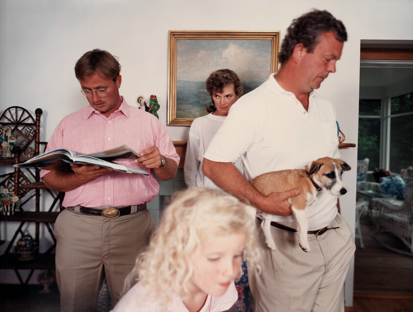 Man carries small dog while another man reads a book and a woman watches behind them.