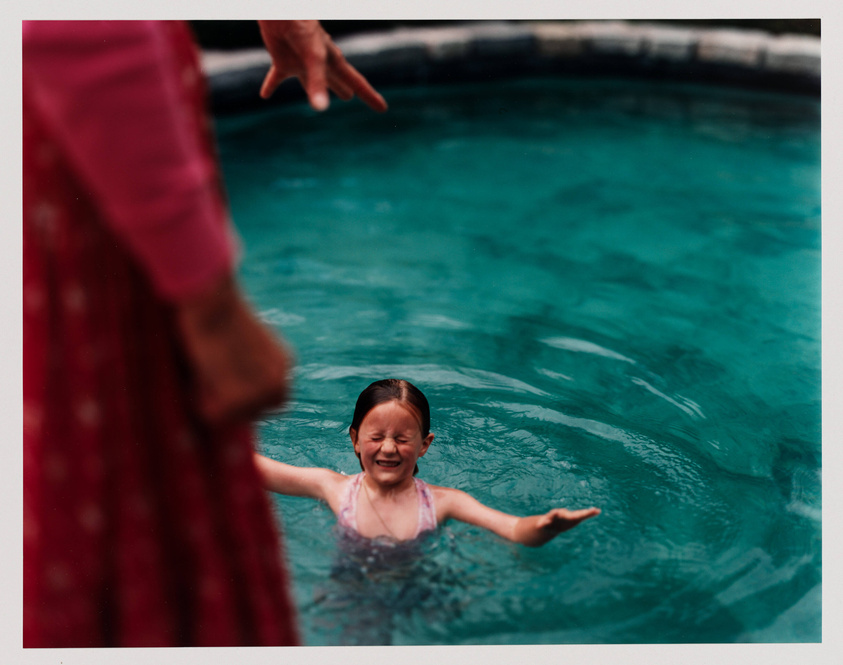 A young child in a pink swimsuit is smiling and reaching out towards an adult's outstretched hand by the edge of a swimming pool, with clear blue water surrounding her.