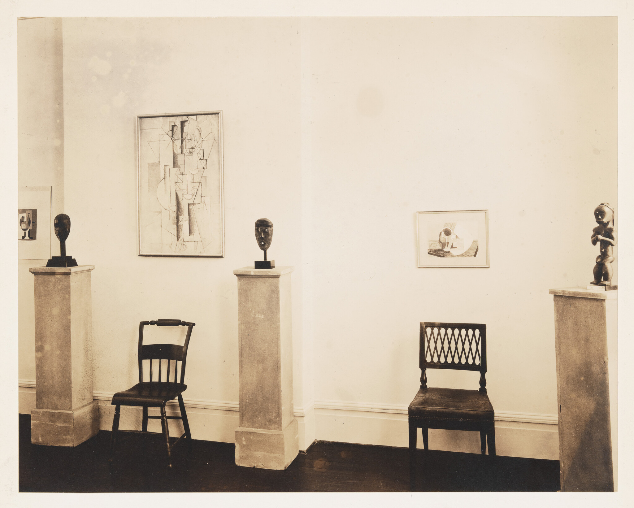 Two wooden chairs sit between pedestals displaying small sculpted heads and a standing figure.