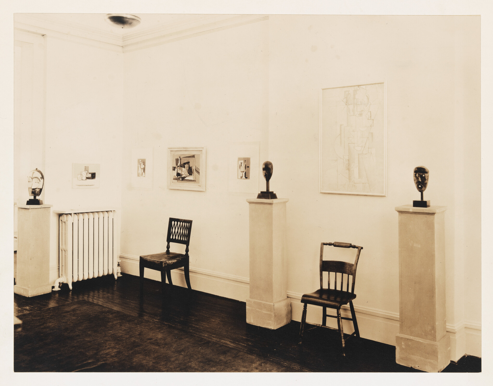 Three sculpted heads on pedestals and two wooden chairs arranged in a quiet gallery room.
