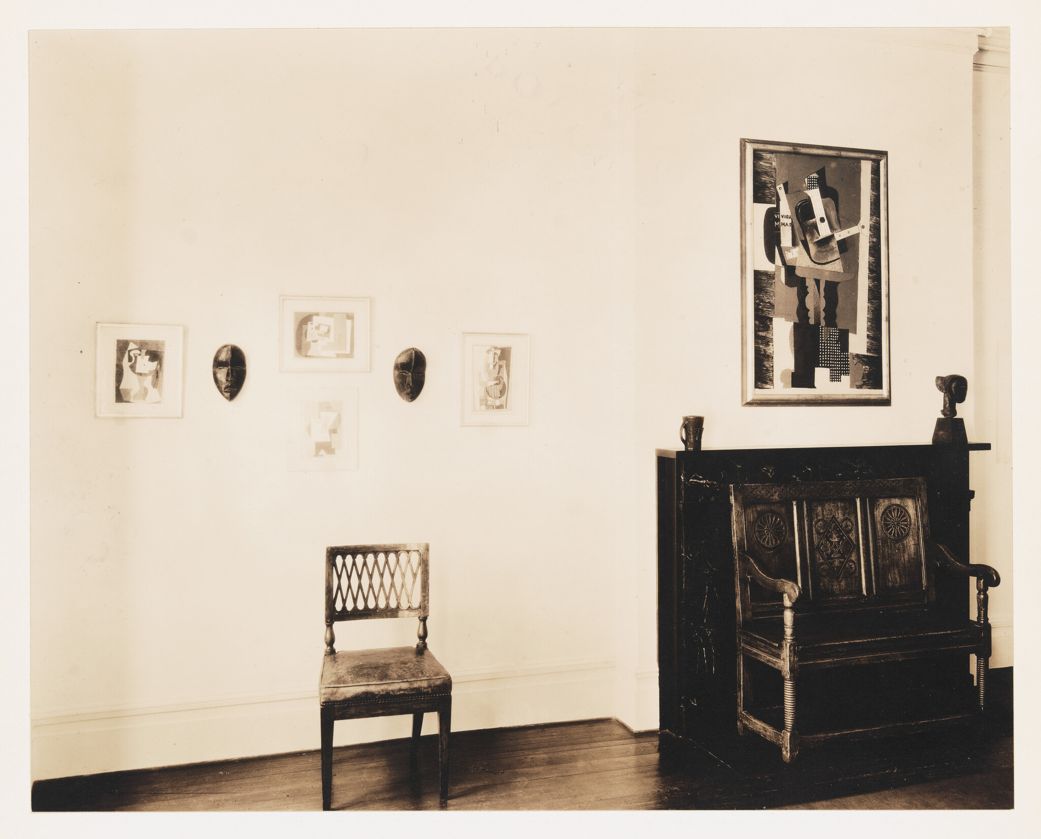 Single wooden chair sits beneath framed abstract prints and two masks beside a dark carved bench.