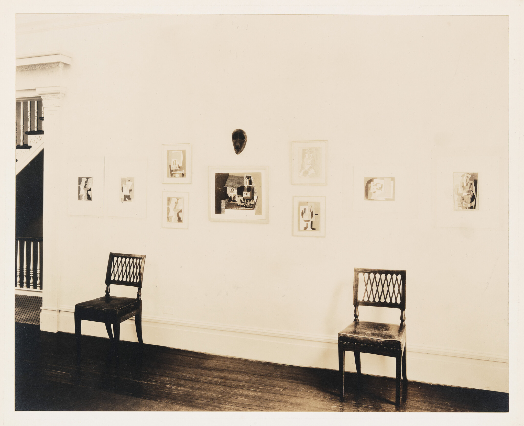 Two wooden chairs face a white wall hung with several framed artworks and a mask.