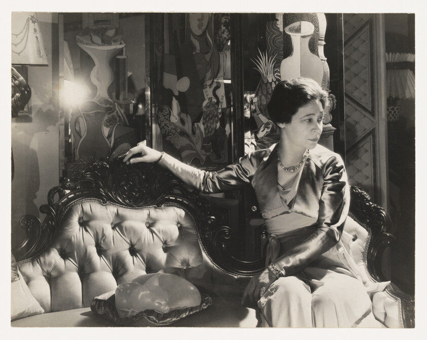 A woman in a satin dress sits on an ornate tufted sofa, looking to the right with hand on backrest.