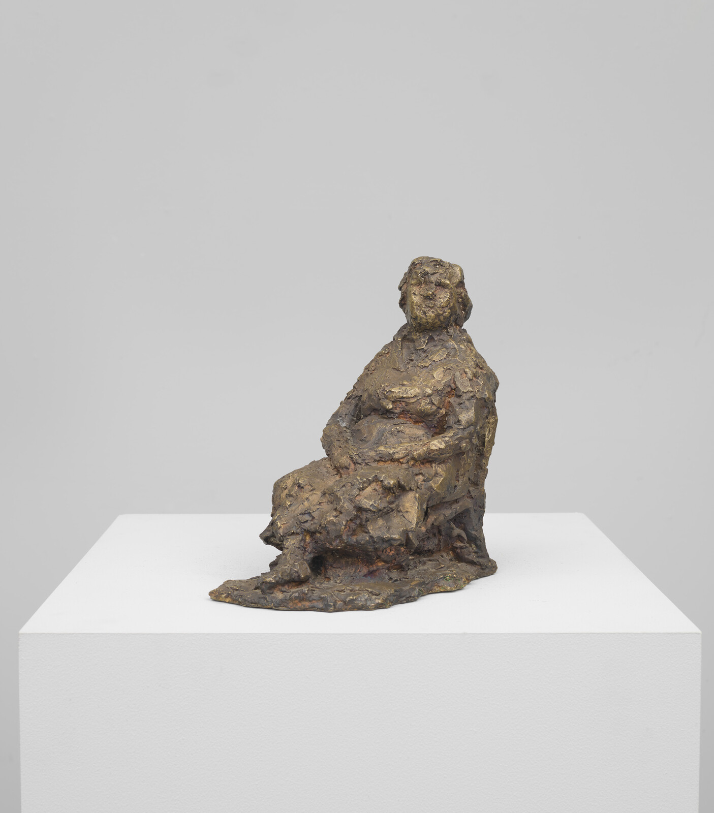 A textured bronze sculpture of a seated figure with an indistinct face, displayed on a white pedestal against a plain background.