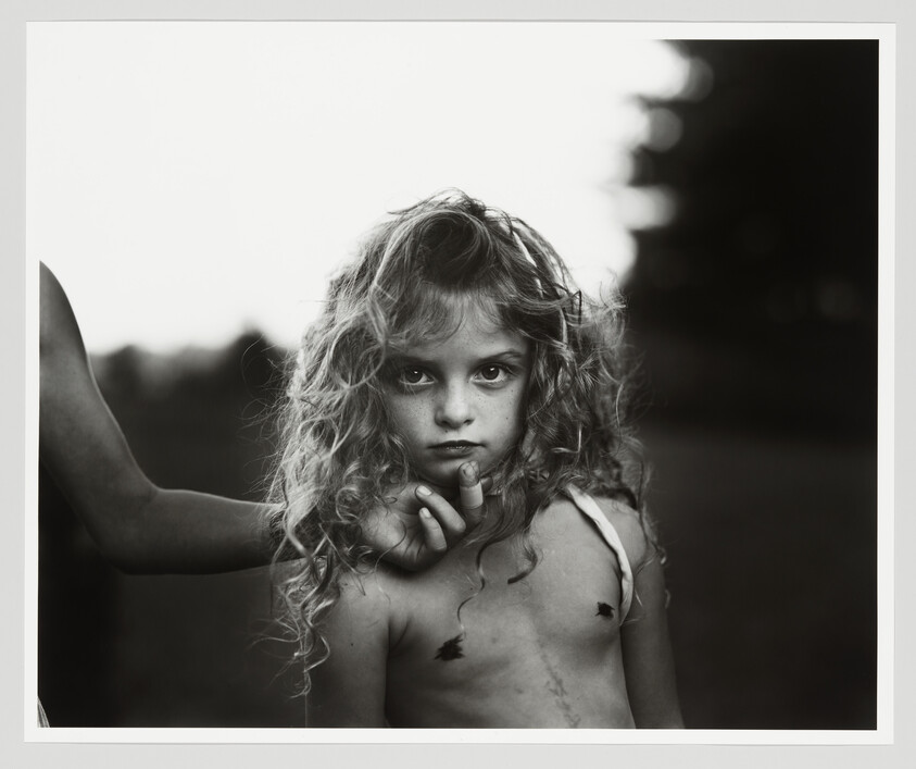 Young girl with curly hair stares at the camera while another person's hand holds her chin.