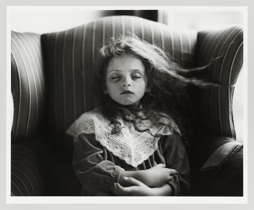 A black and white photograph of a young girl with her eyes closed, appearing to be asleep, sitting in a striped armchair. Her hair is long and wavy, partially covering her face, and she is wearing a vintage dress with a lace collar. The image conveys a sense of peacefulness and nostalgia.