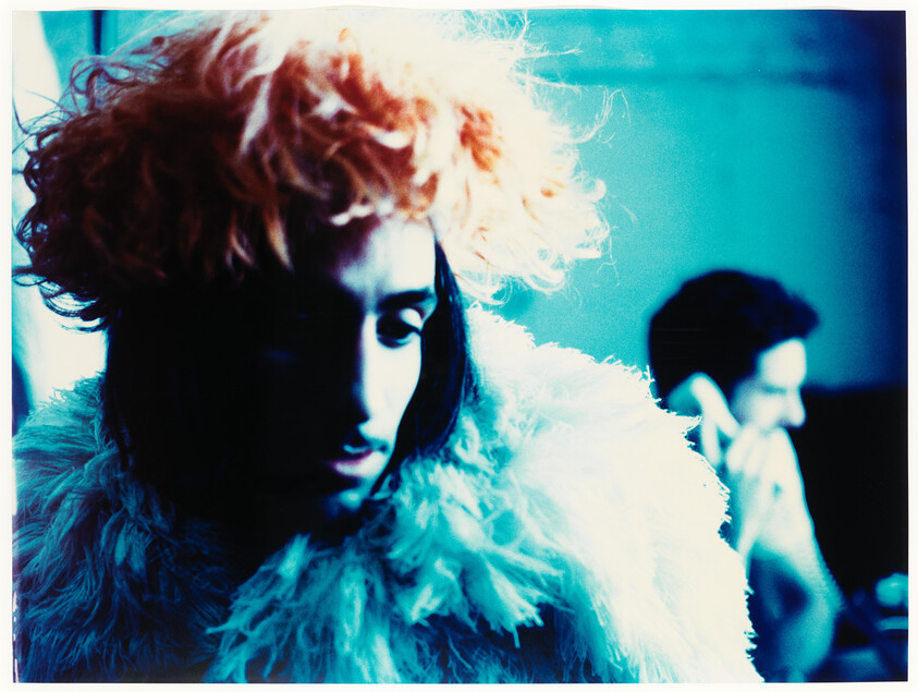 Person wearing a fluffy coat and orange wig looks down while another person talks on a phone.