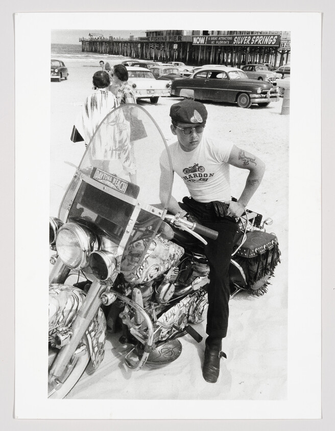 Man wearing a cap sits on a decorated motorcycle on Daytona Beach, with a pier and cars behind.