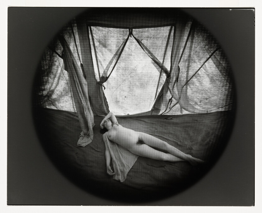 A nude person reclining on a fabric-covered floor inside a tent with draped curtains.