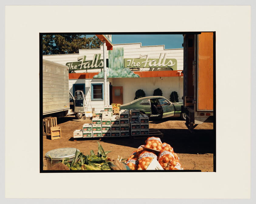 A photograph capturing a sunny day at a roadside establishment named "The Falls," with a mural of a waterfall on the building's facade. In the foreground, there are piles of fresh produce, including onions and corn, with some scattered on the ground. A semi-truck is parked in the middle, partially obscuring the view of an old green car with its door open. The scene suggests a delivery or unloading of goods at this local business.