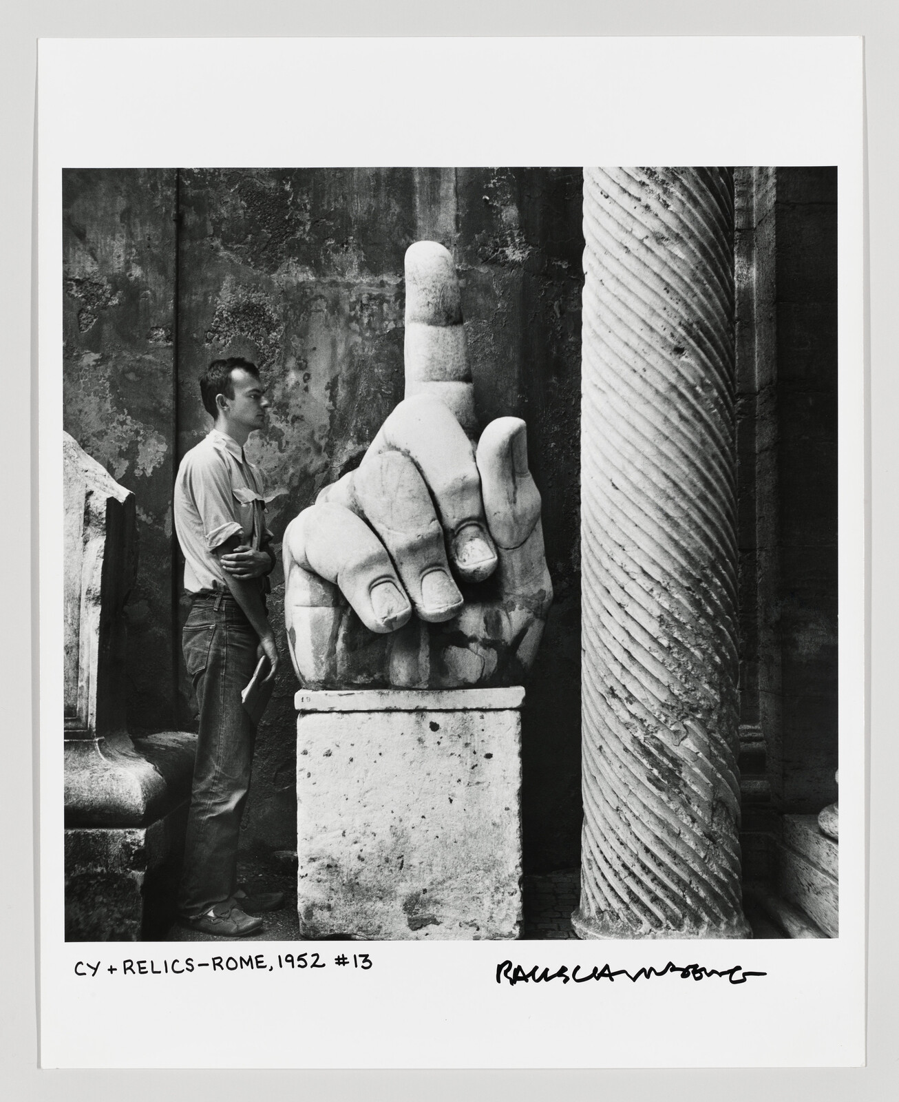 A man stands beside a giant stone hand sculpture with its index finger pointing upward.