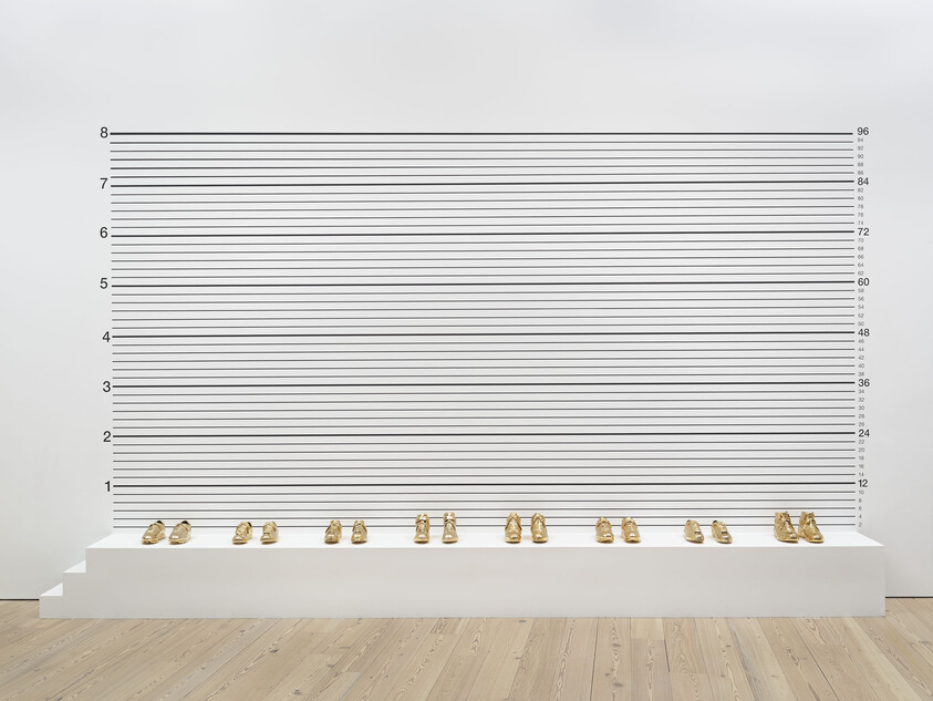 A row of small gold shoes displayed on a white platform against measurement lines.
