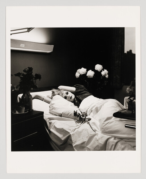A woman lies in a bed surrounded by flowers, holding a rose on the sheets.