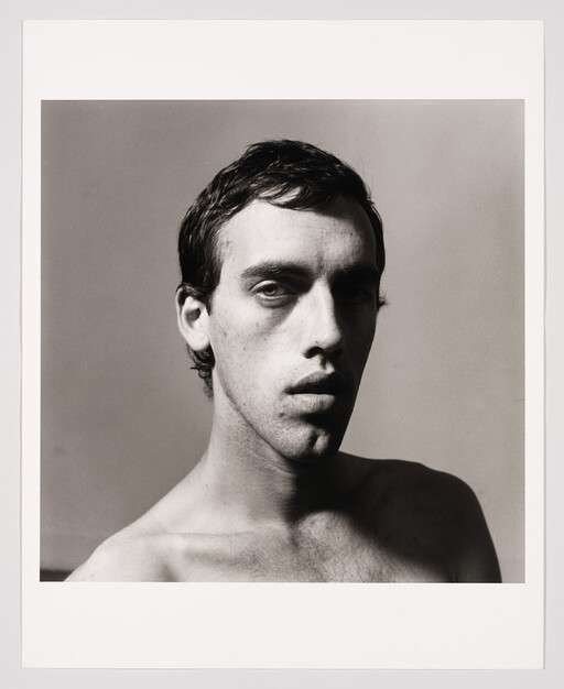 A shirtless young man looks directly at the camera with a neutral, slightly shadowed expression.