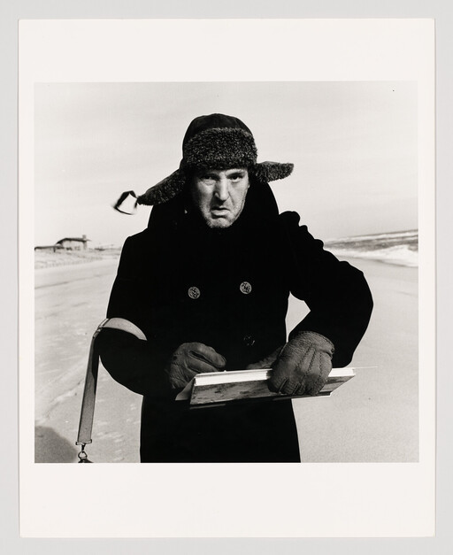 A man in a fur hat and coat writes on a clipboard while standing on a windy beach.