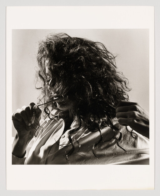 Woman with long curly hair holds a strand in her mouth while another hand pulls hair.